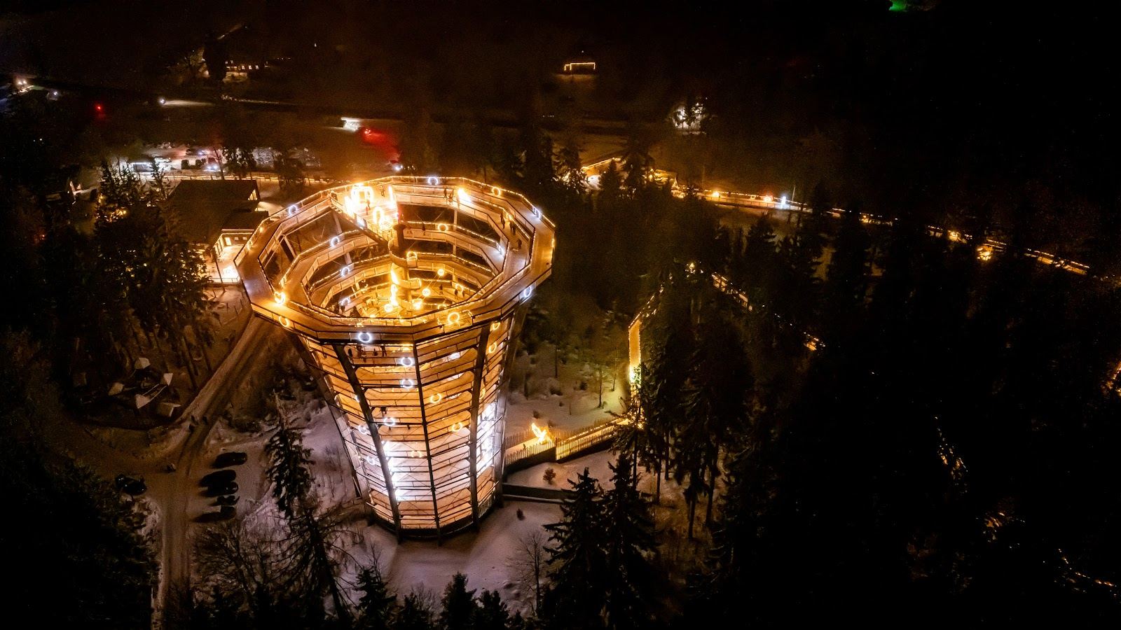 Vista aerea di una torre panoramica illuminata e un sentiero di luci in un bosco innevato.